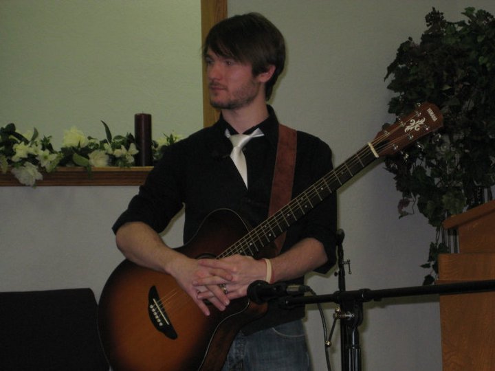 Chris Ulery holding an acoustic guitar in a church with greenery behind