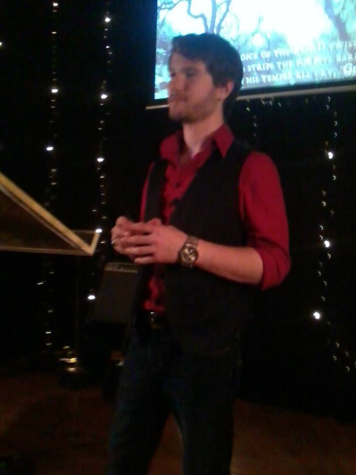 Chris Ulery in a red shirt and vest speaking before string lights