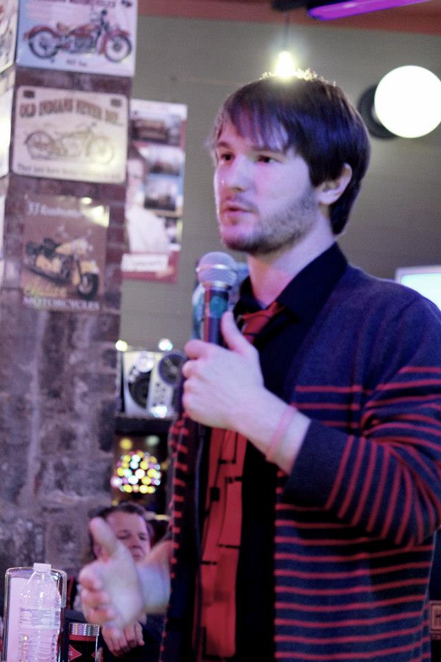 Chris Ulery in a striped cardigan speaking with a microphone and open hand
