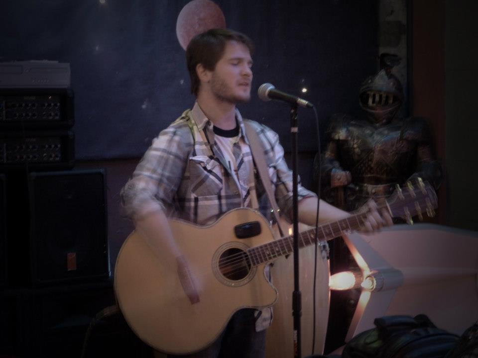 Chris Ulery playing acoustic guitar and singing at a microphone in a dim venue