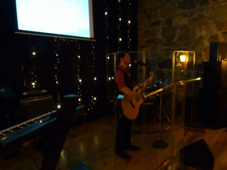 Chris Ulery playing acoustic guitar on a dark stage under string lights