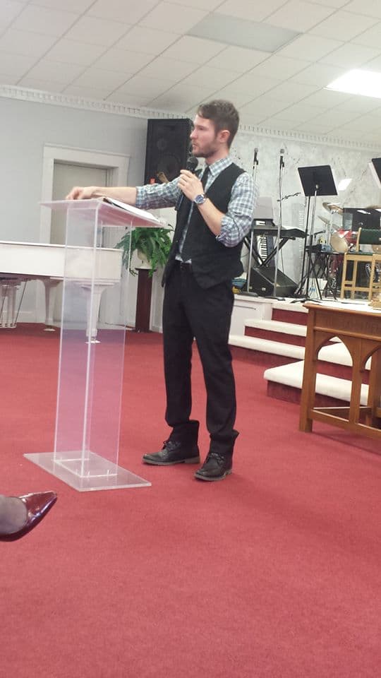 Chris Ulery preaching behind a clear acrylic pulpit in a red-carpeted church sanctuary
