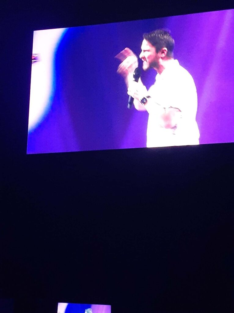 Chris Ulery projected on a giant screen, gesturing toward the crowd while preaching