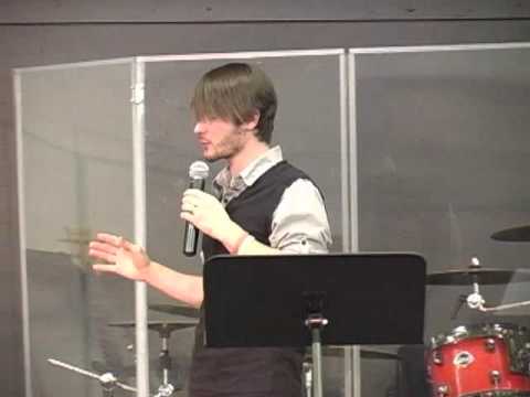 Chris Ulery speaking with a microphone and gesturing near a music stand and drums