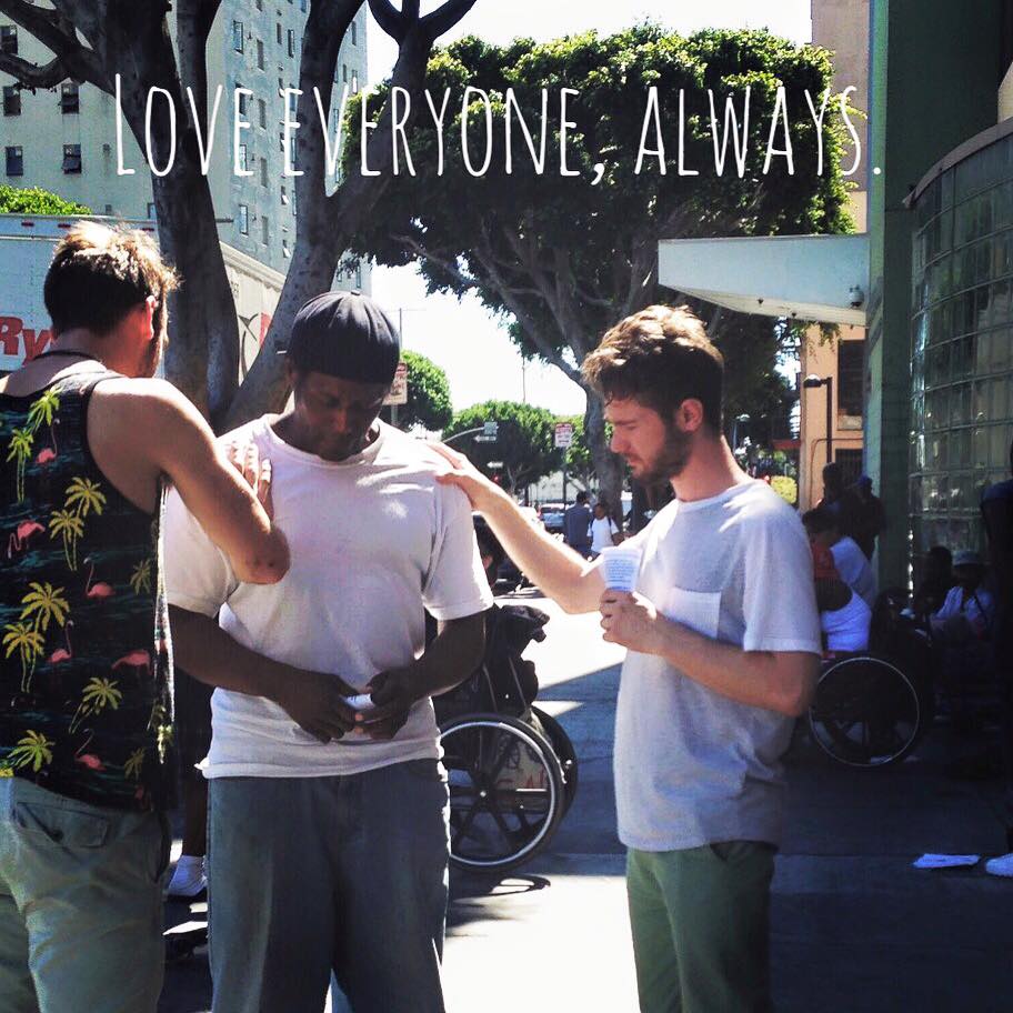 Chris Ulery praying with a man on a city sidewalk during street ministry, with the words Love everyone, always